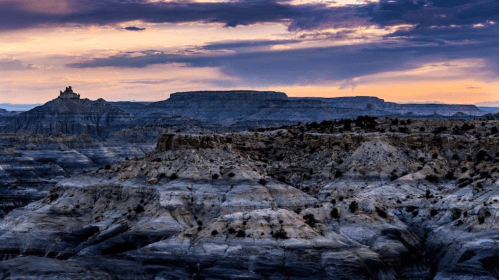 A dramatic landscape featuring layered rock formations and a distant plateau under a colorful sunset sky.
