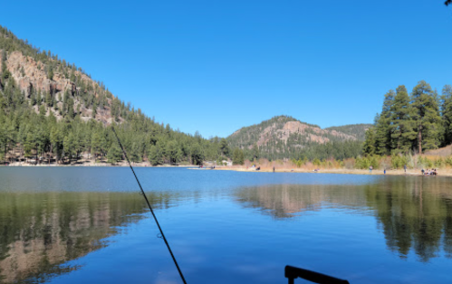 A serene lake surrounded by mountains and pine trees, with a fishing rod in the foreground and people in the distance.