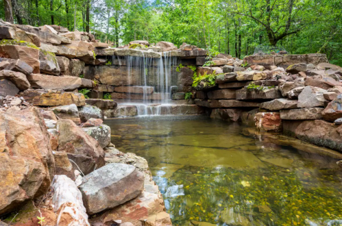 A serene waterfall cascades over rocks into a calm pool, surrounded by lush green trees and natural scenery.