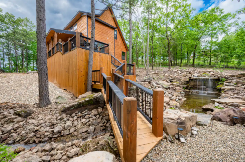 A wooden cabin surrounded by trees, featuring a bridge over a rocky area leading to a small waterfall.