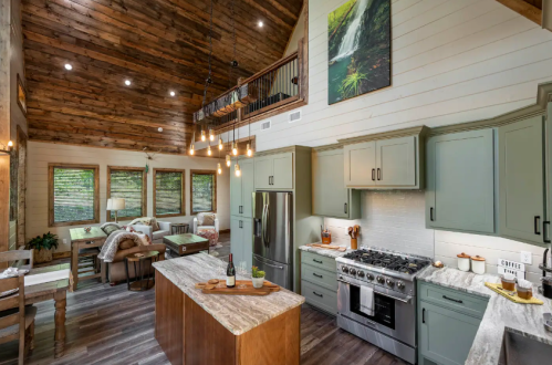 Modern kitchen with green cabinets, marble countertops, and wooden accents, featuring a high ceiling and large windows.
