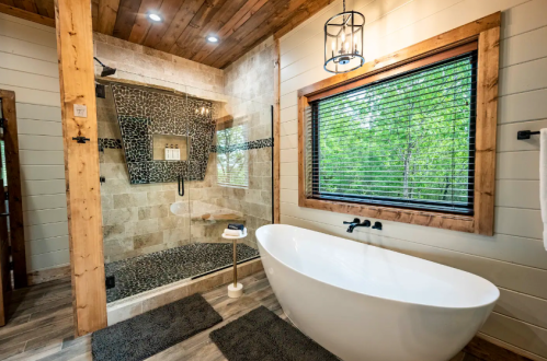 Modern bathroom featuring a walk-in shower, freestanding tub, wooden accents, and large window with greenery outside.