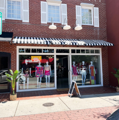A storefront with a striped awning, displaying mannequins in colorful clothing and plants outside.