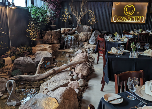 A cozy restaurant interior featuring a rock waterfall, greenery, and elegantly set tables with black tablecloths.