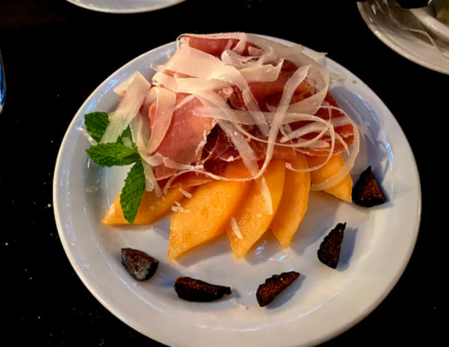 A plate of cantaloupe slices topped with prosciutto, shaved cheese, mint leaves, and garnished with darkened fig pieces.