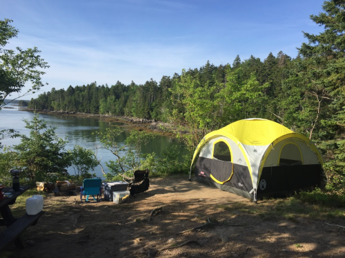 A yellow and black tent by a calm river, surrounded by trees and a sandy area with camping chairs.