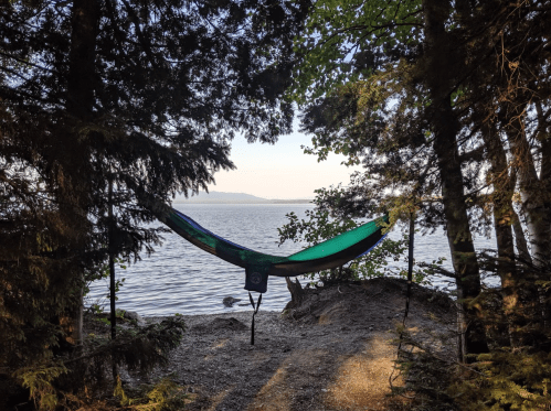 A hammock hangs between trees by a serene lake, surrounded by greenery and a calm water view.