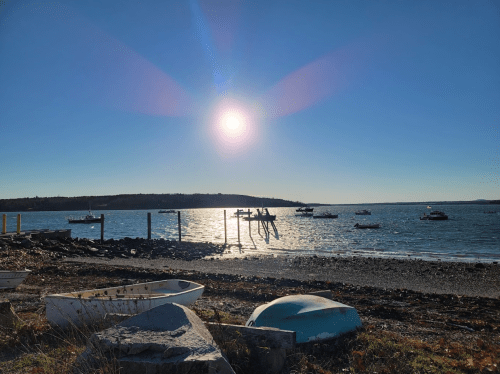 A serene coastal scene with boats on the water, a bright sun, and a clear blue sky reflecting on the waves.