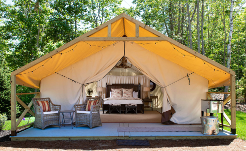 A cozy glamping tent with a bed, seating area, and warm lighting, surrounded by trees.