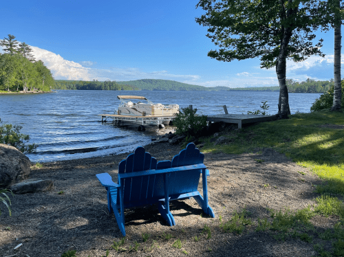 A blue adirondack chair sits on the shore of a lake, with a boat docked nearby and trees in the background.
