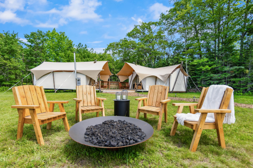 A cozy outdoor campsite with wooden chairs around a fire pit, featuring glamping tents in a lush green setting.