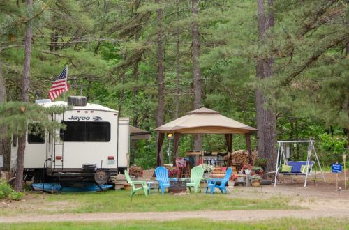 A cozy campsite with a Jayco RV, a gazebo, colorful chairs, and a swing surrounded by tall trees.