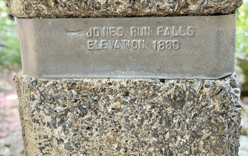 Metal plaque on a stone pillar indicating "Jones Run Falls" and "Elevation 1880." Green foliage in the background.