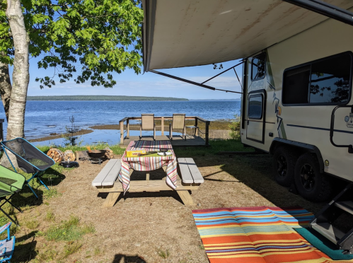 A campsite by the water featuring an RV, picnic table, chairs, and colorful rugs under a shaded awning.