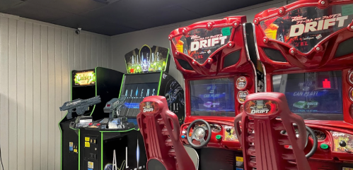A gaming area featuring two red racing arcade machines and a shooting game cabinet in a dimly lit room.
