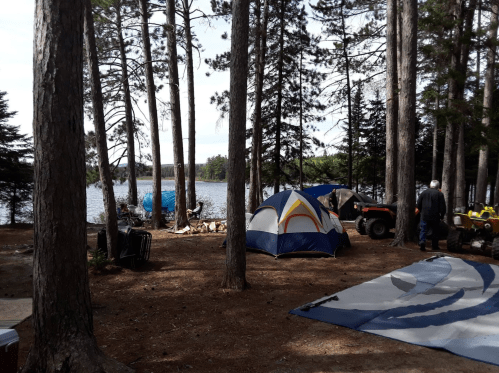 A serene campsite by a lake, featuring tents, trees, and outdoor equipment under a clear sky.