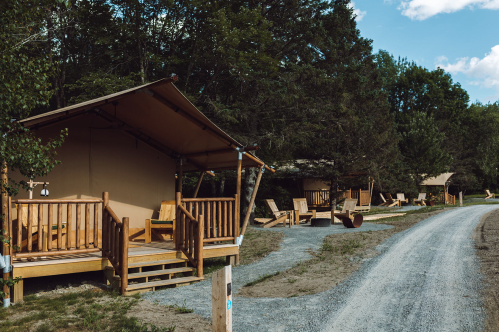 A gravel path leads to wooden glamping tents surrounded by trees, with outdoor seating and a serene atmosphere.