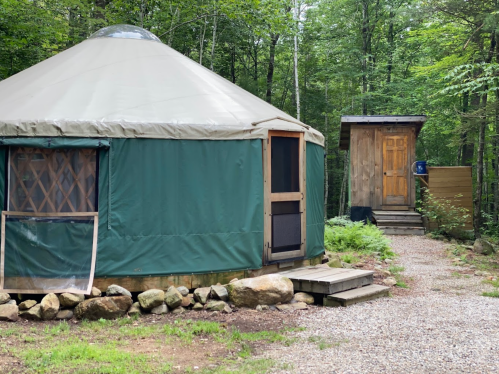 A green yurt with a wooden door, surrounded by trees, next to a small wooden shed along a gravel path.