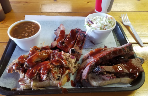 A tray with pulled pork and brisket sandwiches, baked beans, and coleslaw on a wooden table.