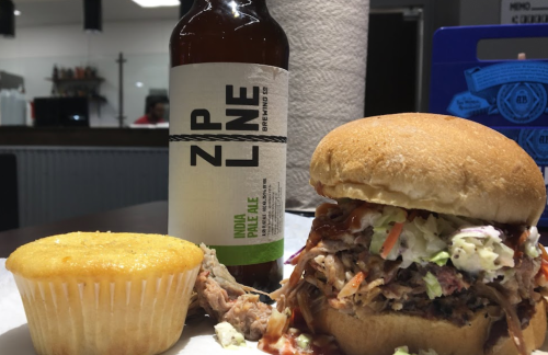 A pulled pork sandwich with coleslaw, a cupcake, and a bottle of ZP LNE India Pale Ale on a table.