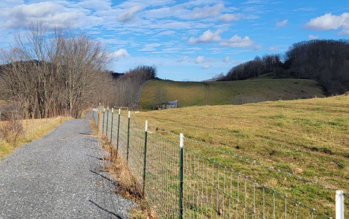 A gravel path lined with a fence leads through a grassy field under a blue sky with scattered clouds.
