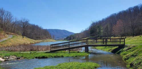 A wooden bridge spans a calm stream, surrounded by green grass and trees, with mountains in the background under a clear blue sky.