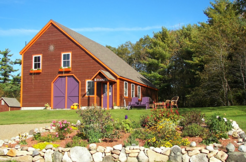 A charming brown barn-style house with purple doors, surrounded by a colorful flower garden and green trees.