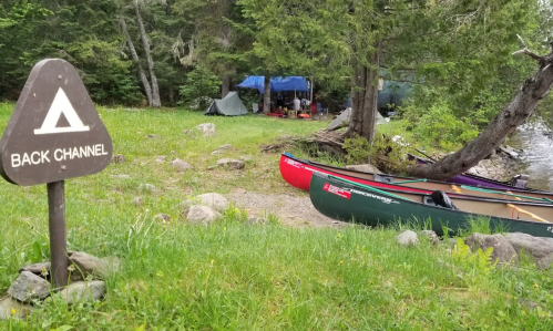 A campsite by a river with a "Back Channel" sign, featuring two canoes and a tent under a blue tarp in the background.