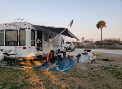 A camper parked on grass with an awning, flags, and outdoor furniture, set against a beach backdrop at sunset.