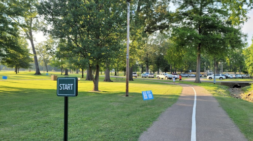 A park path with a "START" sign, surrounded by trees and parked cars in the background.