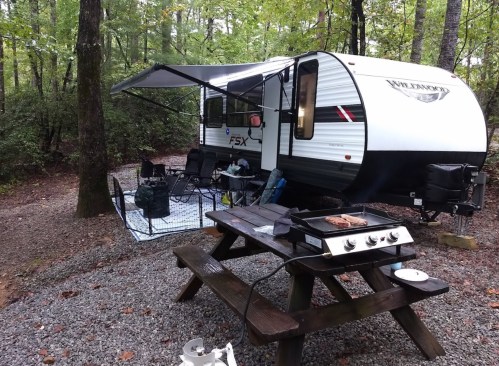 A camping scene featuring a travel trailer under a canopy, with a picnic table and cooking equipment nearby.