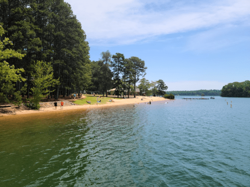 A serene lakeside scene with sandy beach, trees, and people enjoying the water on a sunny day.