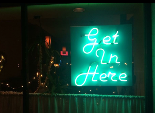 A glowing neon sign that reads "Get In Here" in bright turquoise letters.