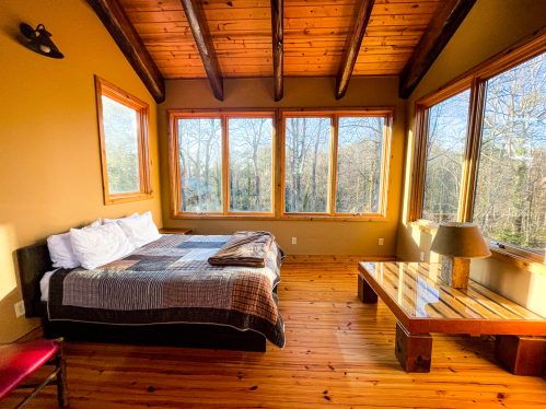 Cozy bedroom with wooden beams, large windows, and a bed with a blanket, overlooking a forested area.