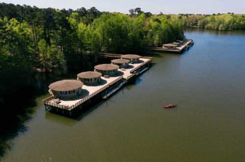 Aerial view of circular cabins on a lake, surrounded by trees, with a kayak paddling nearby.