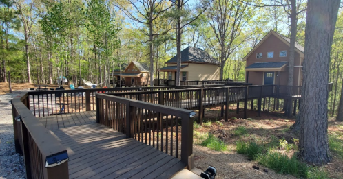 A wooden walkway connects two houses in a wooded area, surrounded by trees and greenery.