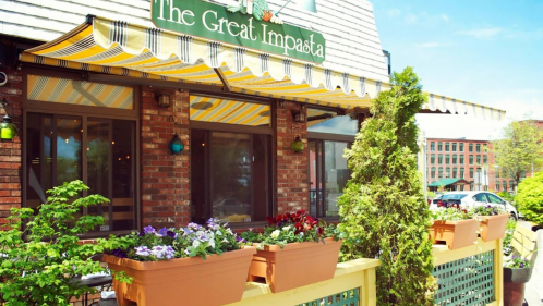 Exterior of The Great Impasta restaurant with colorful flower pots and a striped awning on a sunny day.