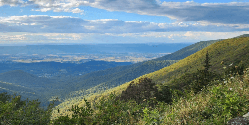 A panoramic view of lush green mountains and valleys under a blue sky with scattered clouds.