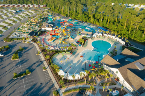 Aerial view of a vibrant water park with slides, pools, and cabanas surrounded by trees and a parking area.