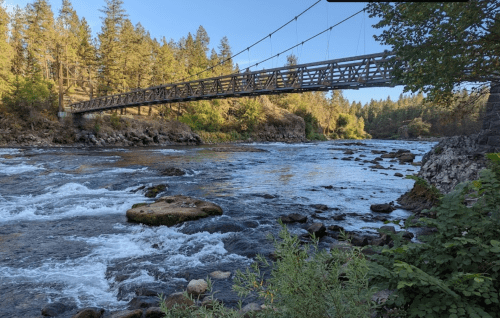 A suspension bridge spans a river surrounded by trees and rocky banks under a clear blue sky.