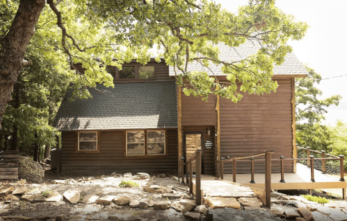 A rustic wooden cabin surrounded by trees, featuring a sloped roof and a welcoming front porch.