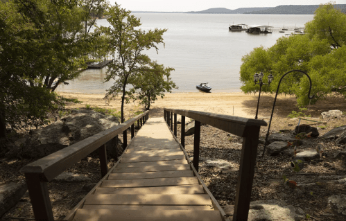 A wooden staircase leads down to a sandy beach by a calm lake, with boats and trees in the background.