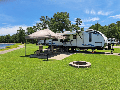 A camper parked by a lake, with a canopy and outdoor seating set up on a grassy area. Trees in the background.