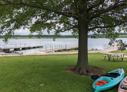 A serene lakeside view with a tree, docks, and kayaks on the grass under a cloudy sky.