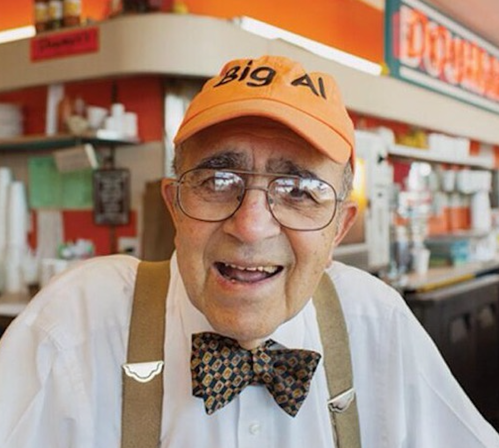 An elderly man with glasses, wearing an orange "Big Al" cap and a bow tie, smiles warmly at the camera in a diner setting.