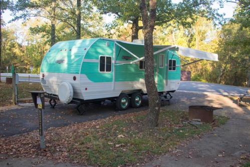 A vintage teal and white camper parked in a wooded area, with a picnic table and fire pit nearby.