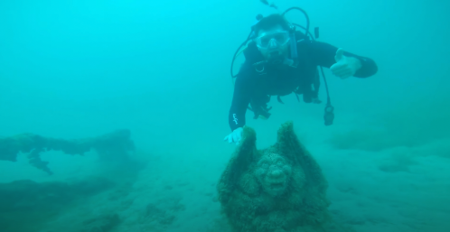 A diver explores an underwater scene with a moss-covered statue resembling a face in a blue, murky environment.