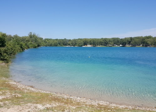 A serene lake with clear blue water, surrounded by green trees and a sandy shore under a clear blue sky.