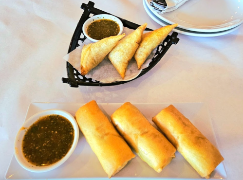 A plate of golden spring rolls and samosas with dipping sauces on a white tablecloth.