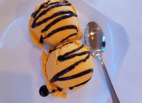 Two scoops of orange ice cream drizzled with chocolate sauce on a white plate, accompanied by a silver spoon.